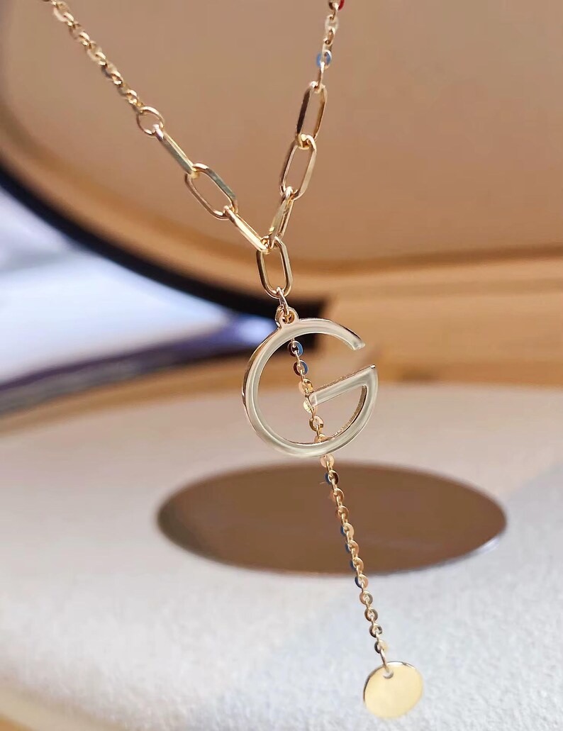 Puede incluir: Collar dorado con un gran colgante con la letra "G". El collar tiene una cadena delicada con un peque&ntilde;o dije circular. El dise&ntilde;o incluye diferentes estilos de cadena, creando un aspecto en capas. La joya se muestra sobre un fondo suave.