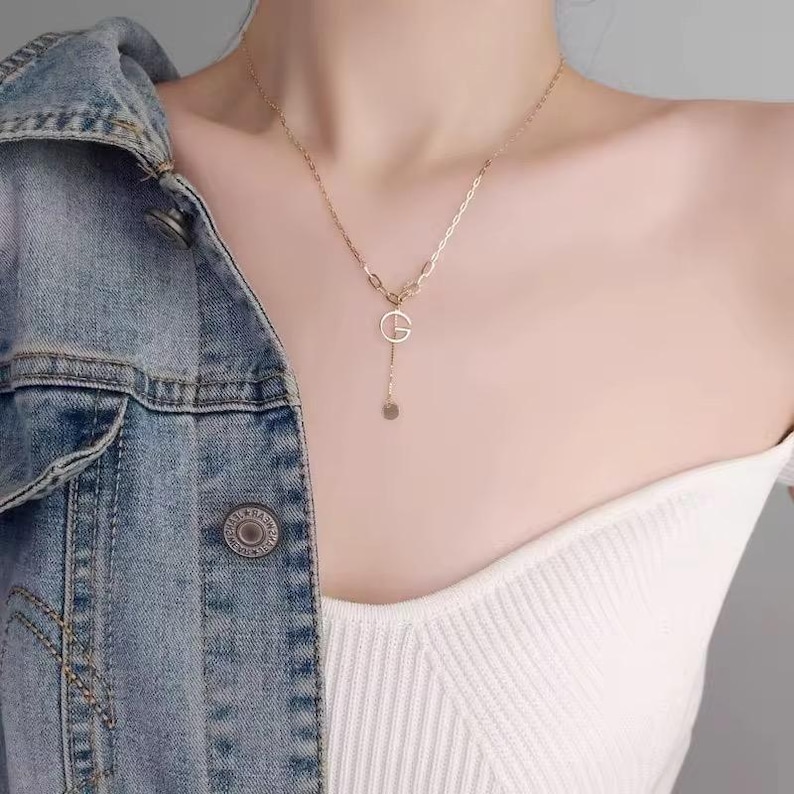 Puede incluir: Un collar dorado con un dise&ntilde;o de eslabones y un colgante con la letra "G" y un peque&ntilde;o disco. El collar se lleva sobre una camiseta blanca y una chaqueta vaquera.