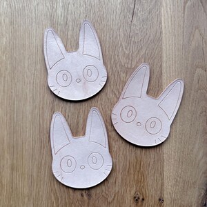 May include: Three light brown cat-shaped wooden coasters. Each coaster features a detailed outline of a cat's face with large eyes, a small nose, and pointed ears. The coasters are arranged on a wooden surface.