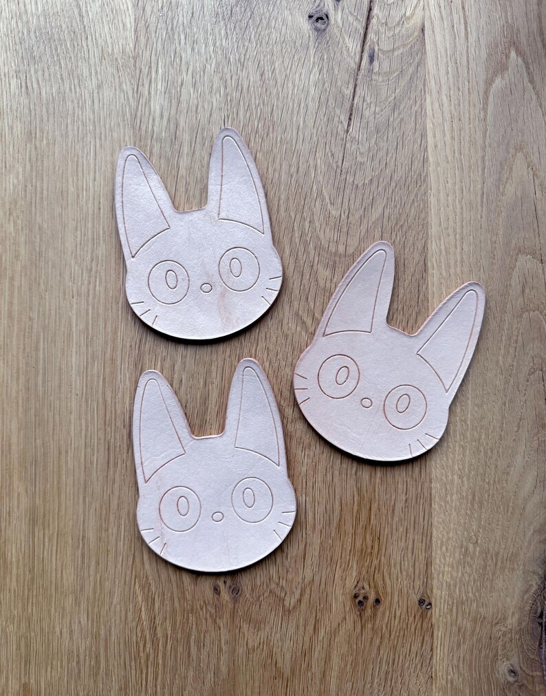May include: Three light brown cat-shaped wooden coasters. Each coaster features a detailed outline of a cat's face with large eyes, a small nose, and pointed ears. The coasters are arranged on a wooden surface.