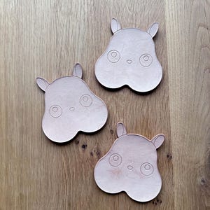 May include: Three light pink hippo-shaped coasters with etched details, including eyes, nostrils, and ears. The coasters are arranged on a wooden surface, showcasing their unique design and color.