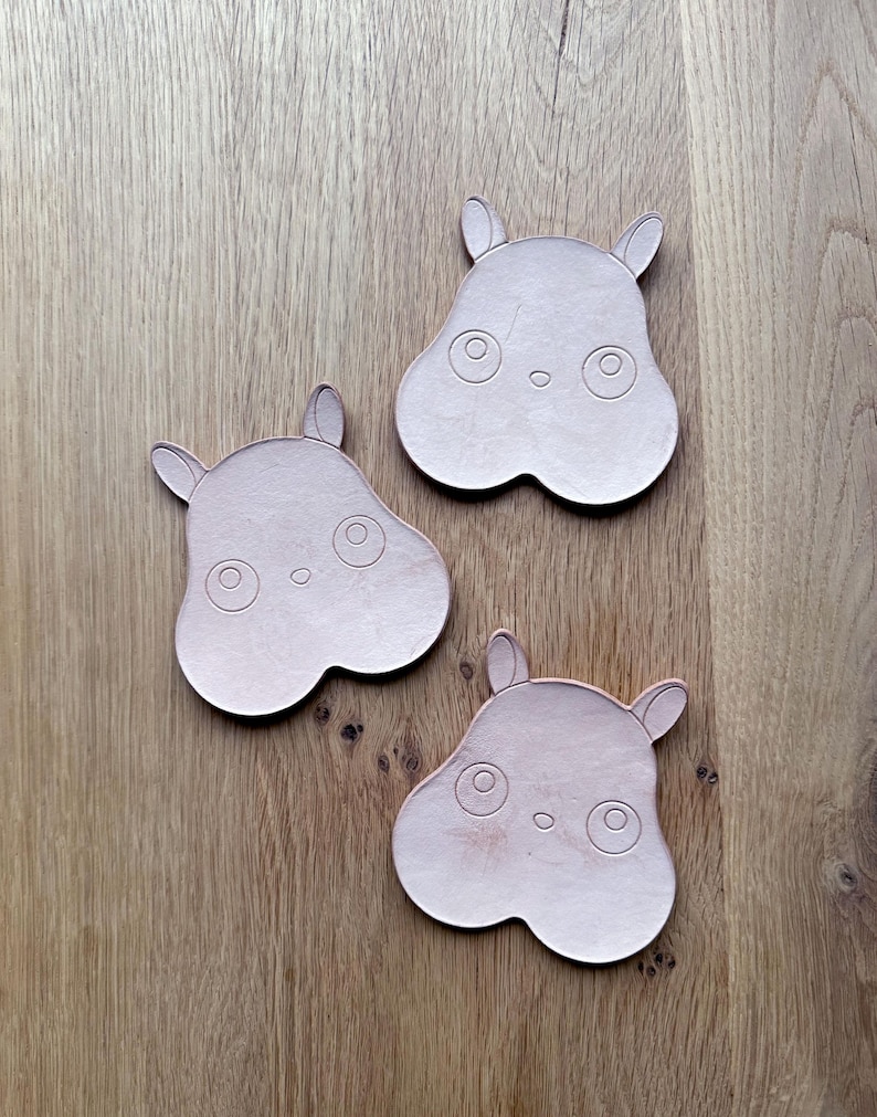 May include: Three light pink hippo-shaped coasters with etched details, including eyes, nostrils, and ears. The coasters are arranged on a wooden surface, showcasing their unique design and color.