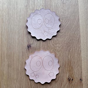 May include: Two wooden coasters with a cartoon eye design and star accents. The coasters have a scalloped edge and are light brown in color. They are placed on a wooden surface.