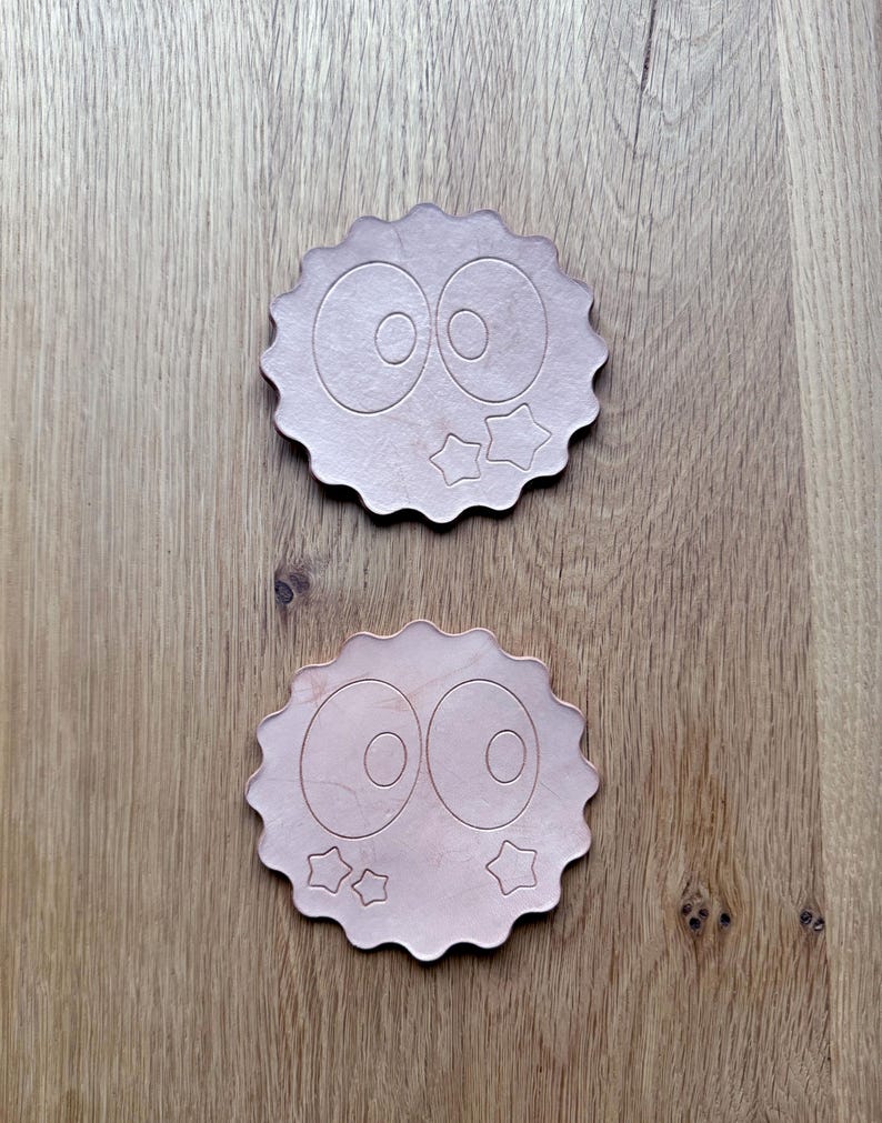 May include: Two wooden coasters with a cartoon eye design and star accents. The coasters have a scalloped edge and are light brown in color. They are placed on a wooden surface.
