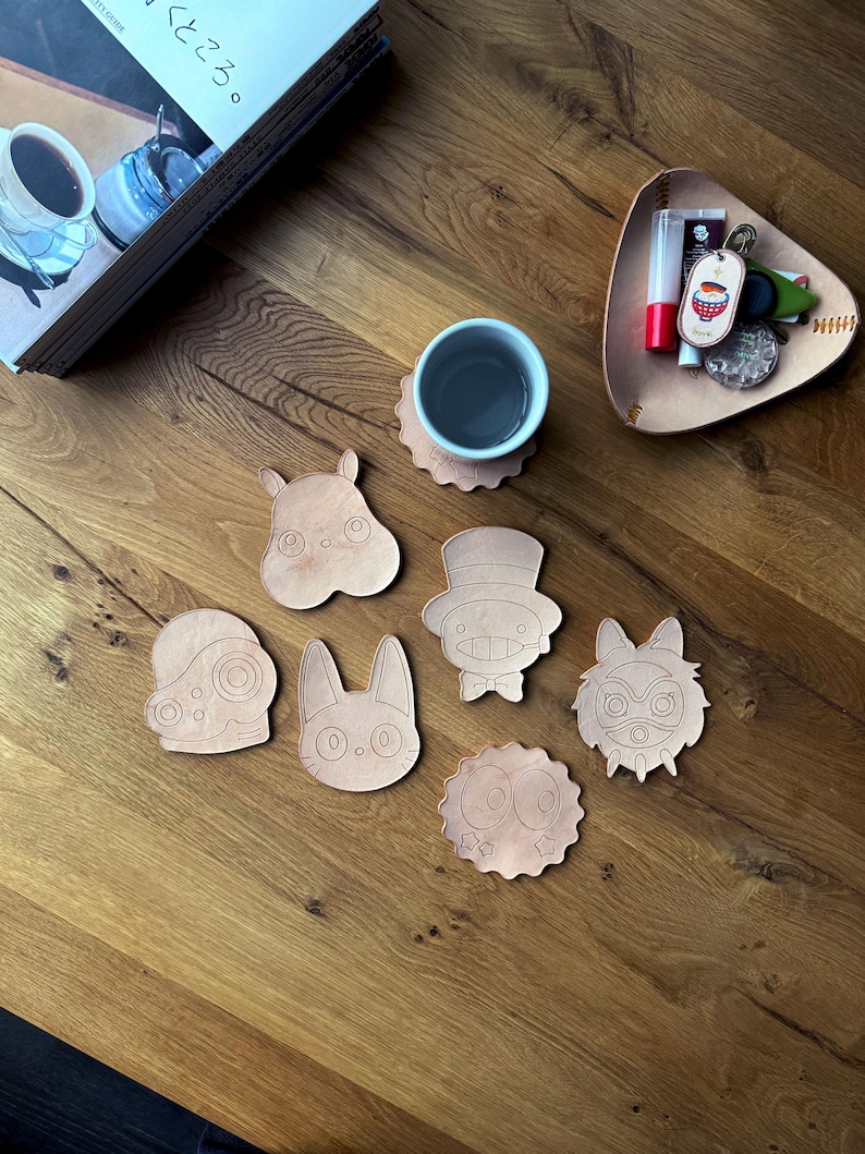 May include: A collection of light brown leather coasters in various cartoon character shapes, including a hippo, rabbit, and owl. A cup of tea sits on a matching coaster. A small leather tray holds keys and lip balm.