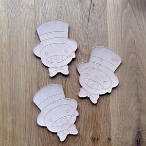 May include: Three light brown, die-cut leather coasters. Each coaster features a cartoon character with a top hat, bow tie, and a smiling face. The coasters are arranged on a wooden surface.