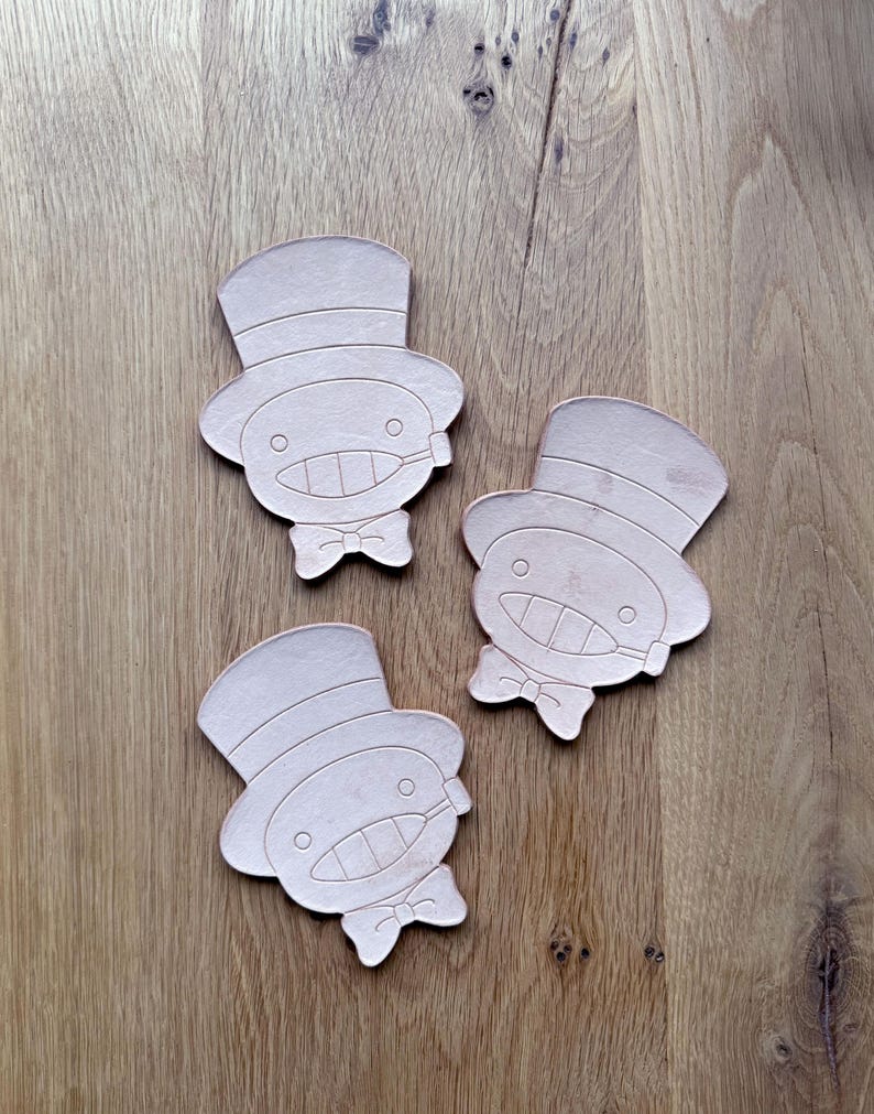 May include: Three light brown, die-cut leather coasters. Each coaster features a cartoon character with a top hat, bow tie, and a smiling face. The coasters are arranged on a wooden surface.