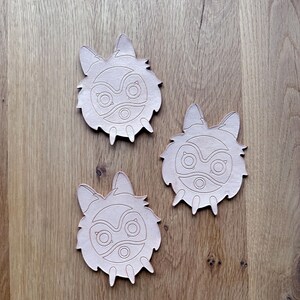 May include: Three light brown leather coasters with a stylized wolf or fox face design. Each coaster features a detailed cutout of the face, including eyes, nose, and ears. The coasters are arranged on a wooden surface.
