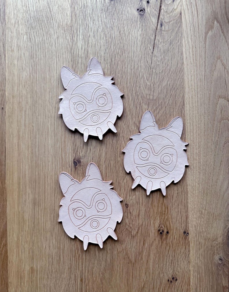 May include: Three light brown leather coasters with a stylized wolf or fox face design. Each coaster features a detailed cutout of the face, including eyes, nose, and ears. The coasters are arranged on a wooden surface.