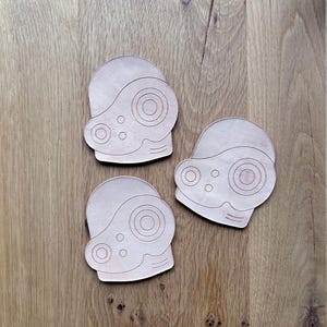May include: Three light brown leather coasters, each shaped like a stylized helmet with circular and oval cutouts. The coasters are arranged on a wooden surface, showcasing their design and texture.