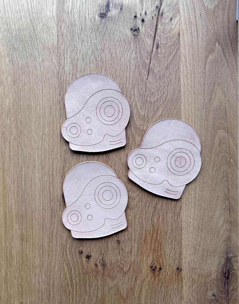May include: Three light brown leather coasters, each shaped like a stylized helmet with circular and oval cutouts. The coasters are arranged on a wooden surface, showcasing their design and texture.