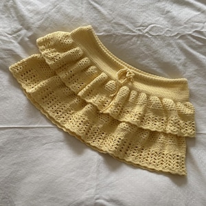 以下が含まれることがあります： 明るい黄色の手作りのかぎ針編みスカート。ティアードフリルデザイン。スカートは透かし模様とドローストリングクロージャーが特徴です。柔らかいパステルカラーと繊細な質感は、夏の衣服を示唆しています。