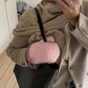 Peut inclure: Un mitaine rose clair, en crochet &agrave; motif d'&eacute;toiles, avec un petit d&eacute;tail pompon. La mitaine est tenue dans un sac fourre-tout noir &agrave; motif de losanges. La personne porte un blazer et une &eacute;charpe beiges.