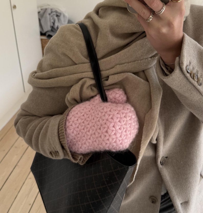 Peut inclure: Un mitaine rose clair, en crochet &agrave; motif d'&eacute;toiles, avec un petit d&eacute;tail pompon. La mitaine est tenue dans un sac fourre-tout noir &agrave; motif de losanges. La personne porte un blazer et une &eacute;charpe beiges.