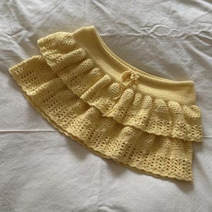 Könnte beinhalten: Hellgelber, gehäkelter Rock mit einem mehrschichtigen, gerüschten Design. Der Rock hat ein Lochmuster und einen Kordelzug in der Taille. Der handgefertigte Rock ist aus weichem Garn.