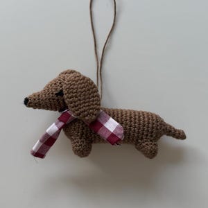 Könnte beinhalten: Gehäkelter Dackel-Ornament in Braun mit einem rot-weiß karierten Schal. Der Hund hat eine kleine schwarze Nase und wird an einer braunen Schnur aufgehängt. Ein handgefertigtes Dekorationsstück.