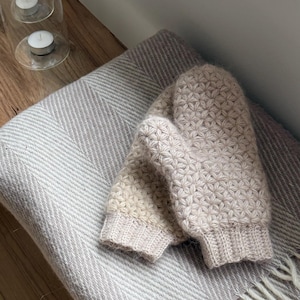Peut inclure: Une paire de mitaines en tricot beige clair avec un motif floral, pos&eacute;e sur une couverture &agrave; chevrons grise et blanche claire avec des franges. Un bougeoir en verre avec deux bougies allum&eacute;es est en arri&egrave;re-plan.