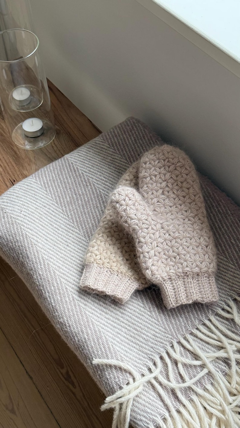 Peut inclure: Une paire de mitaines en tricot beige clair avec un motif floral, pos&eacute;e sur une couverture &agrave; chevrons grise et blanche claire avec des franges. Un bougeoir en verre avec deux bougies allum&eacute;es est en arri&egrave;re-plan.