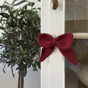 May include: A knitted burgundy bow hangs from a white cabinet door. The bow, made of textured yarn, has a classic shape with two loops and two long tails. A bronze knob is visible above the bow. The cabinet is next to a plant.