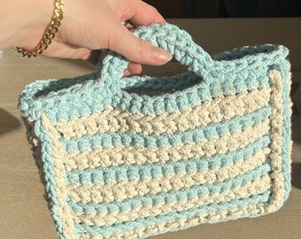 May include: A small, crocheted tote bag with a light blue and cream striped pattern. The bag has a single handle and is made with a textured stitch.