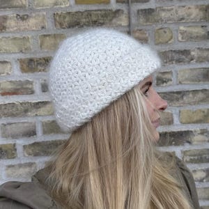 Puede incluir: Un gorro blanco tejido con un patrón texturizado en forma de estrella. El gorro está hecho de un material suave y esponjoso. El fondo es una pared de ladrillos. La persona lleva un gorro y tiene el pelo largo y rubio.