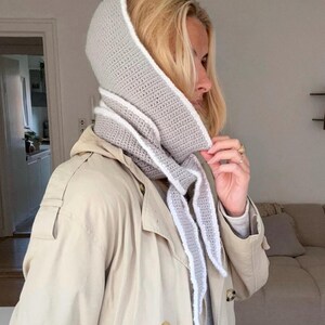 Op de afbeelding: Een lichtgrijze gehaakte capuchon met witte rand en lange sjaalachtige uiteinden. De capuchon wordt over een lichtgekleurde trenchcoat gedragen. De capuchon is een stijlvol accessoire voor koeler weer.