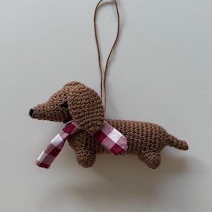 Puede incluir: Adorno de perro salchicha marrón tejido a ganchillo con una bufanda a cuadros rojos y blancos. El perro tiene un cuerpo largo, patas cortas y una cola pequeña. Una cuerda marrón está unida para colgar.