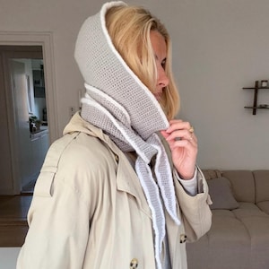 Op de afbeelding: Een lichtgrijze gehaakte capuchon met een witte rand en lange, puntige uiteinden. De capuchon wordt over een beige trenchcoat gedragen. Een stijlvol accessoire voor koeler weer.