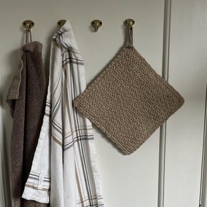 Puede incluir: Tres toallas de cocina colgadas de ganchos en una puerta blanca. Las toallas son una toalla de rizo marrón, una toalla a cuadros blancos y marrones y un agarrador de olla de ganchillo marrón.