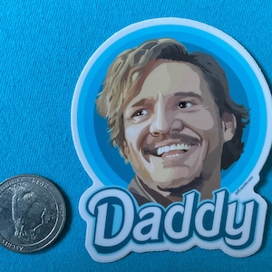 May include: A white sticker featuring a cartoon illustration of a smiling man with light brown hair and a mustache, set against a blue background. The word "Daddy" is written in a playful font below the image. A coin is next to the sticker.