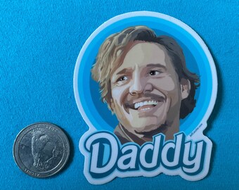 May include: A white sticker with a cartoon illustration of a smiling man with light brown hair and a mustache, set against a blue background. The word "Daddy" is written in a playful font below the image. A quarter is next to the sticker.