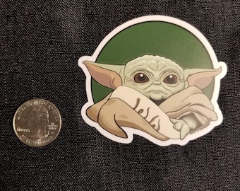 May include: A green and brown cartoon sticker of a character from the Star Wars franchise, known as Baby Yoda, or The Child. The character is depicted with large eyes and a serious expression, wearing a brown robe and sitting with its arms crossed.