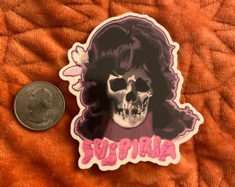Puede incluir: Una pegatina con una calavera con pelo oscuro y una flor, con un suéter morado. La pegatina tiene la palabra "SUSPIRIA" en letras rosas con burbujas. Un cuarto de dólar estadounidense está al lado de la pegatina.