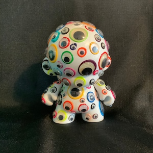 May include: White toy figure with a round head covered in colorful googly eyes. The figure has arms and legs and is standing on a black surface.