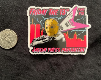 May include: A rectangular sticker featuring a yellow hockey mask-wearing figure, a pink electric guitar, and a red New York City skyline silhouette. The text reads FRIDAY THE 13TH PART VIII JASON TAKES MANHATTAN. A US quarter is shown for scale.