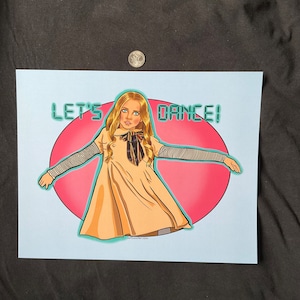May include: A digital illustration of a girl with long blonde hair wearing a beige dress and a black and gold bow tie. She is standing with her arms outstretched and the text "LET'S DANCE!" is written in a teal green font above her head. The background is a pink circle.