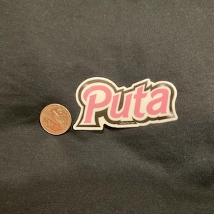 May include: A sticker with the word "Puta" in pink, outlined in black and white, with a coin next to it for scale. The sticker has a glossy finish and is on a black surface.