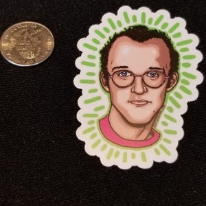May include: A sticker featuring a cartoon portrait of a man with brown hair, wearing glasses and a pink shirt. The background is green with a sunburst design.