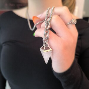 May include: A silver-toned chain necklace with a geometric rose quartz crystal pendant. The pendant is a light pink color and is held by a silver metal frame. The necklace is being held up by a person wearing a black top.