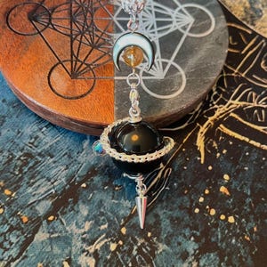 May include: A silver pendant with a black orb, silver wire detail, and a silver cone dangle. It includes a crescent moon with a yellow gemstone and a silver beaded sphere. The background shows a wooden coaster with geometric designs.