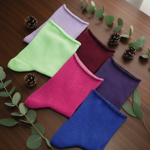 May include: An assortment of colorful socks in shades of purple, green, pink, burgundy, blue, and purple. The socks are arranged on a wooden surface with pine cones and eucalyptus leaves.