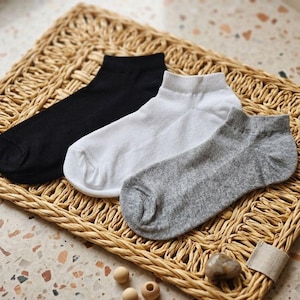 May include: Three pairs of ankle socks in black, white, and grey are displayed on a woven wicker tray. The socks are made of a soft material and are neatly folded. The background is a speckled surface.
