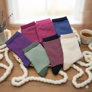 Può includere: Una collezione di calzini a righe colorati in varie tonalità di viola, rosa, blu, verde e beige. I calzini sono disposti in un cestino intrecciato su una superficie di legno, con una tazza di tè ed elementi decorativi sullo sfondo.
