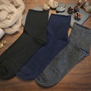 May include: Three pairs of socks in black, navy blue, and gray are displayed on a wooden surface. The socks are folded and arranged side by side. Additional items include yarn, twine, buttons, and decorative stones.