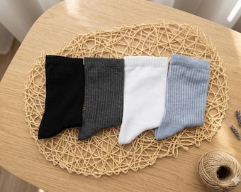 May include: Four pairs of socks in black, dark gray, white, and light blue with vertical ribbed texture. The socks are arranged on a woven placemat, with a ball of twine and lavender sprigs nearby. The socks are made of a soft material.
