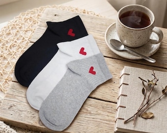 May include: Three pairs of socks in black, white, and gray, each with a small red heart embroidered on the cuff. The socks are arranged on a wooden tray next to a cup of tea and a small notebook. A cozy, neutral-toned blanket is in the foreground.