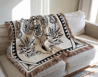 May include: A black and white woven throw blanket with a tiger head, roses, and rope design. The blanket is fringed and has a cream background.