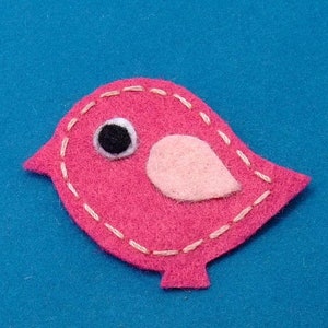 Puede incluir: Un pájaro de fieltro rosa con un ala de fieltro rosa y un ojo negro. El pájaro está cosido con hilo blanco alrededor de los bordes.