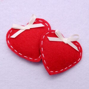 May include: Two red felt hearts with white satin bows. The hearts are stitched with white thread and have a white background.