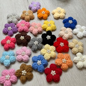 May include: A collection of 25 crocheted flower embellishments in various colors, including pink, purple, blue, red, yellow, orange, brown, and gray. Each flower has a center of white pearl beads.